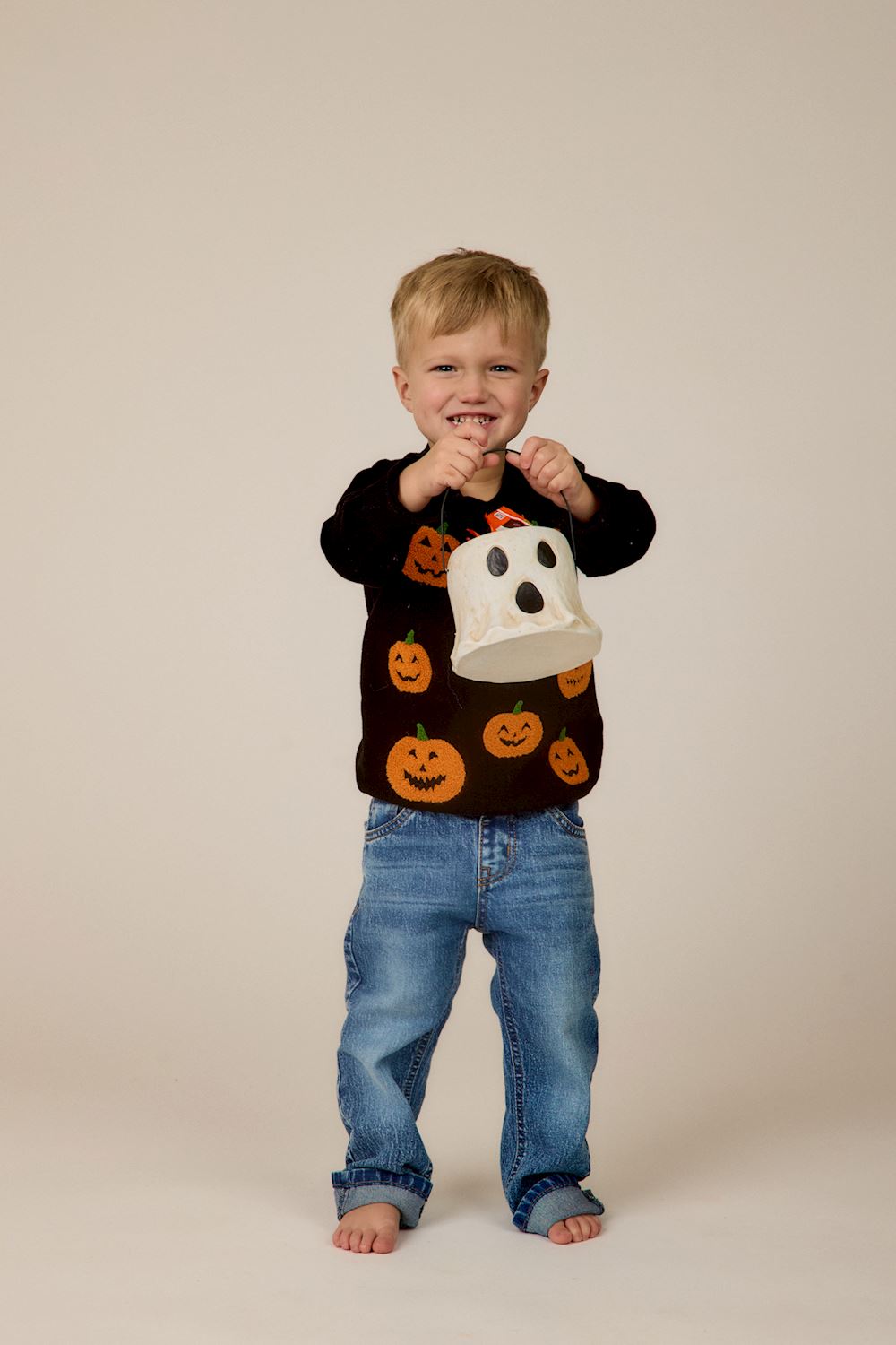 A young toddler boy wearing a black long-sleeve sweater with orange pumpkin prints and blue jeans, holding a white ghost-shaped candy bucket