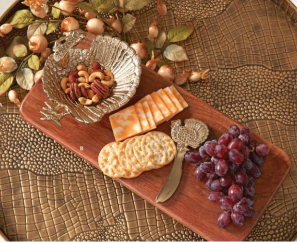 A wooden charcuterie board styled with crackers, cheese, grapes, nuts, and a decorative cheese knife with a turkey top and an attached turkey dish