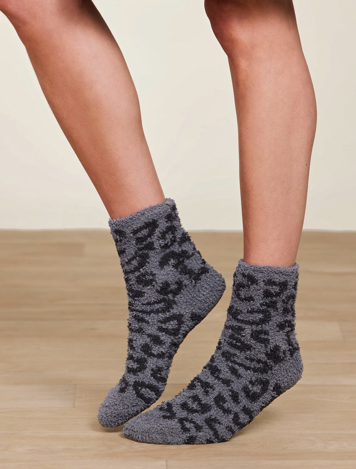 Gray fuzzy socks with black leopard print pattern worn by a person on a wooden floor.