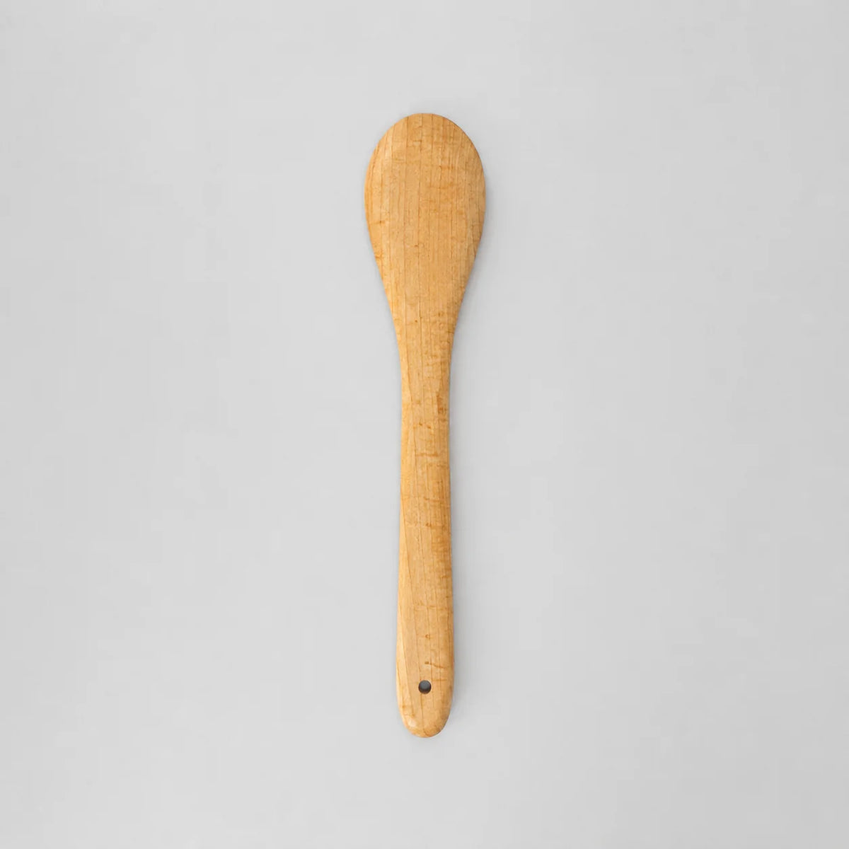 A light natural maple wood spoon with a small metal rivet at the end of the handle, by holland bowl mill shown vertically against a plain light gray background.