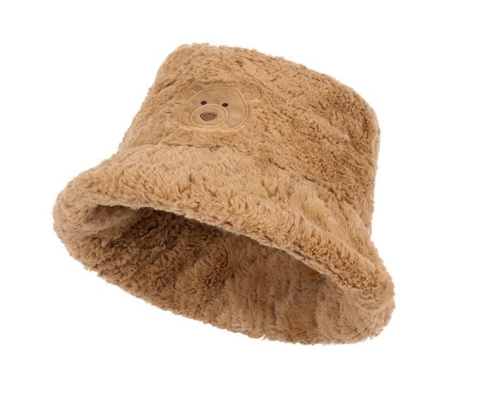A plush tan bucket hat with a teddy bear face patch on the front, shown against a plain white background.