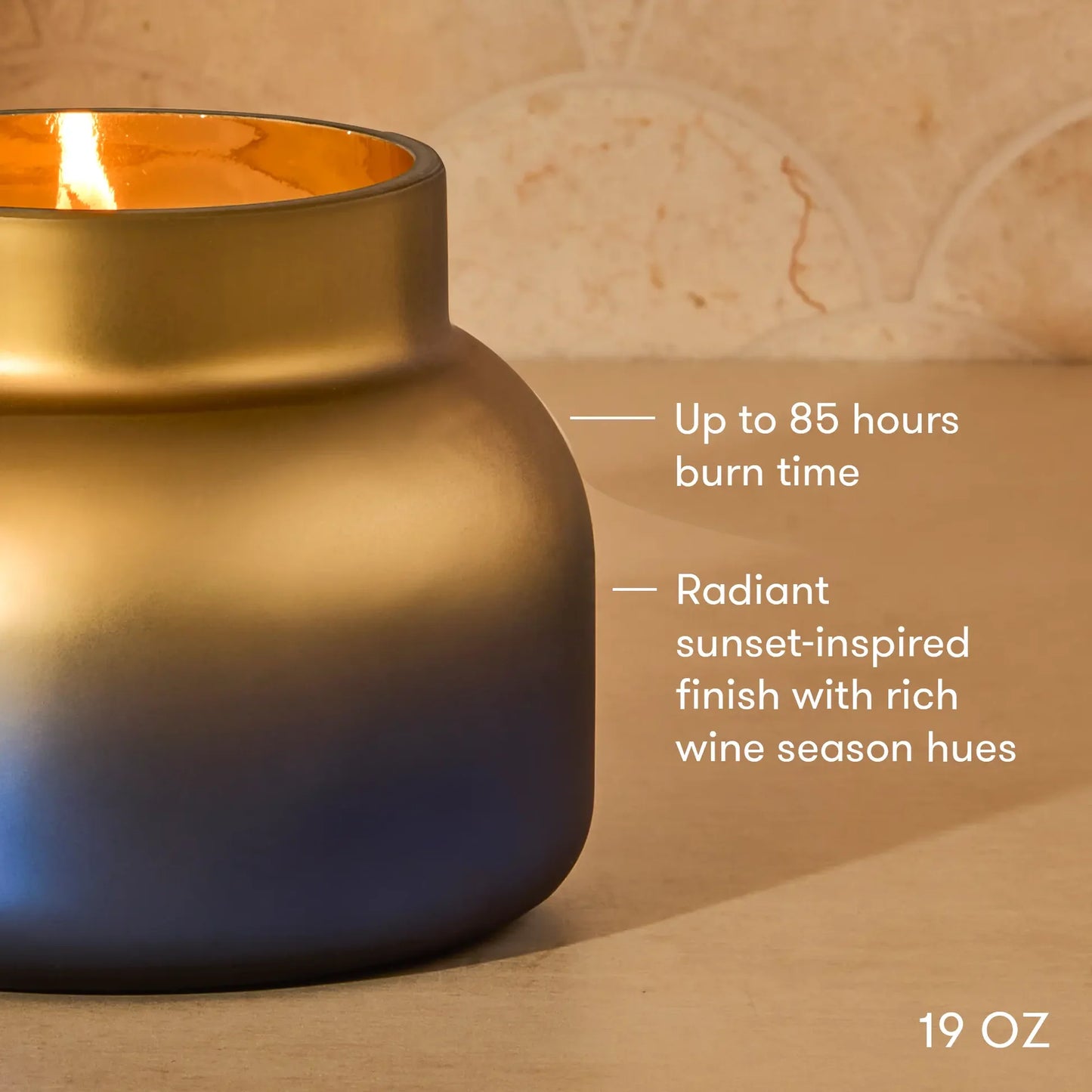 A 19 oz candle in a matte-finish vessel with a gradient color shift from gold at the top to deep blue at the bottom, lit with a warm flame. White text on the right highlights its 85-hour burn time and sunset-inspired wine season hue.