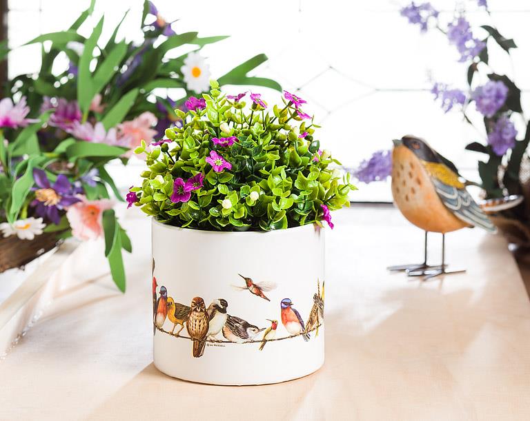 A white ceramic planter decorated with colorful bird illustrations, filled with green artificial foliage and small pink flowers, set on a tabletop with blurred flowers and a bird figurine in the background.