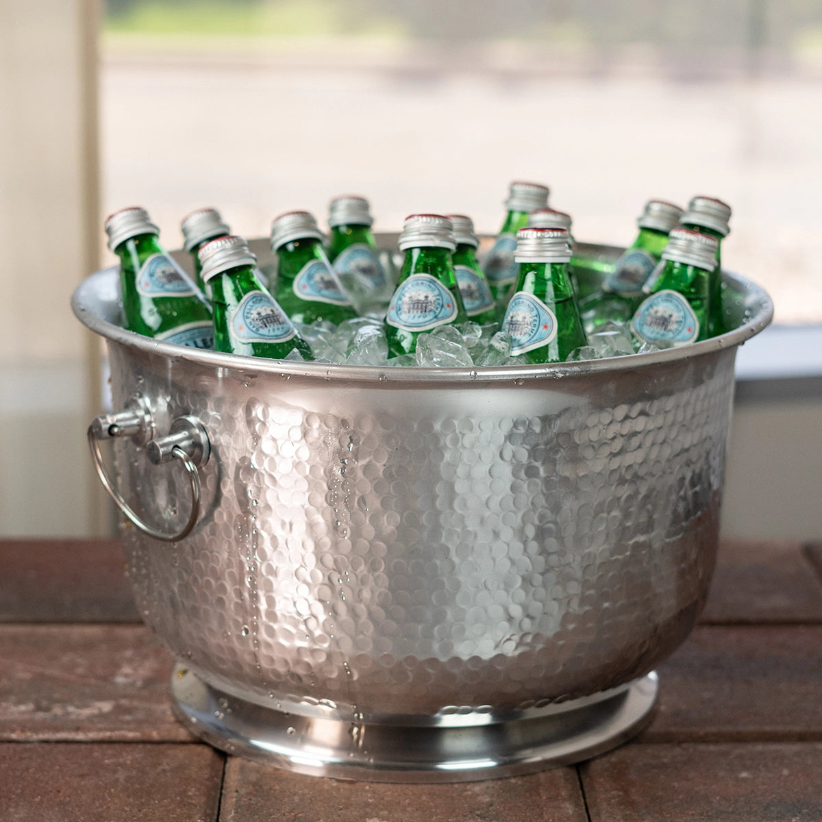 Silver Metal ice bucket with 2 handles filled with green bottles on a wooden surface