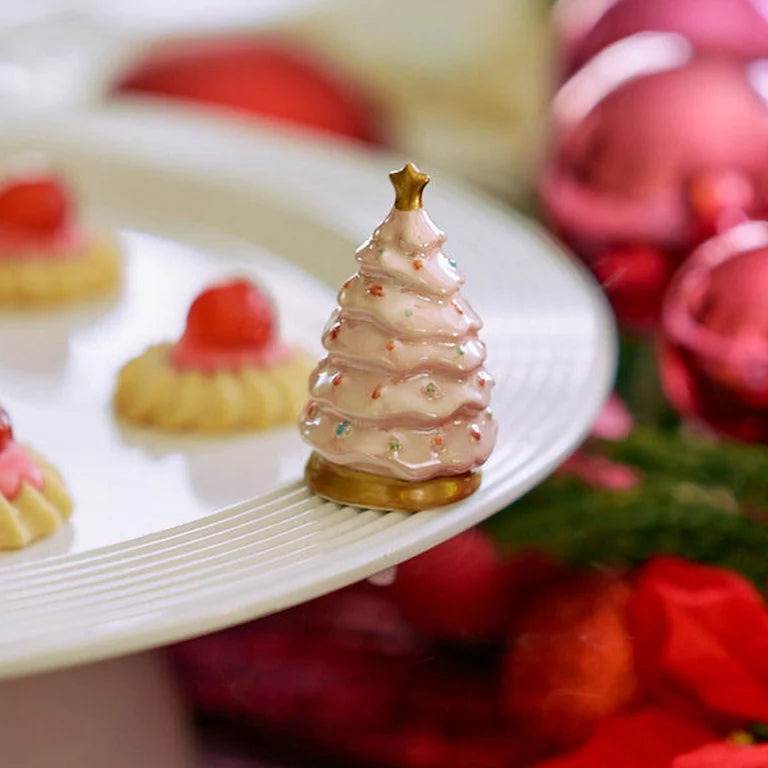 A small pink frosted Christmas tree decoration with a gold star topper, displayed on a white plate with other decorated pastries and red holiday ornaments in the background.