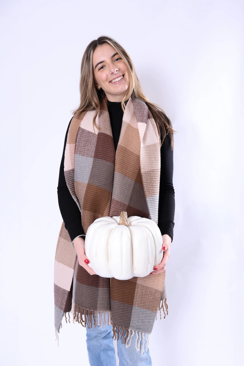 A woman modeling a large plaid fringed wrap scarf in autumn shades of brown, beige, and gray, holding a white decorative pumpkin against a plain white studio background.