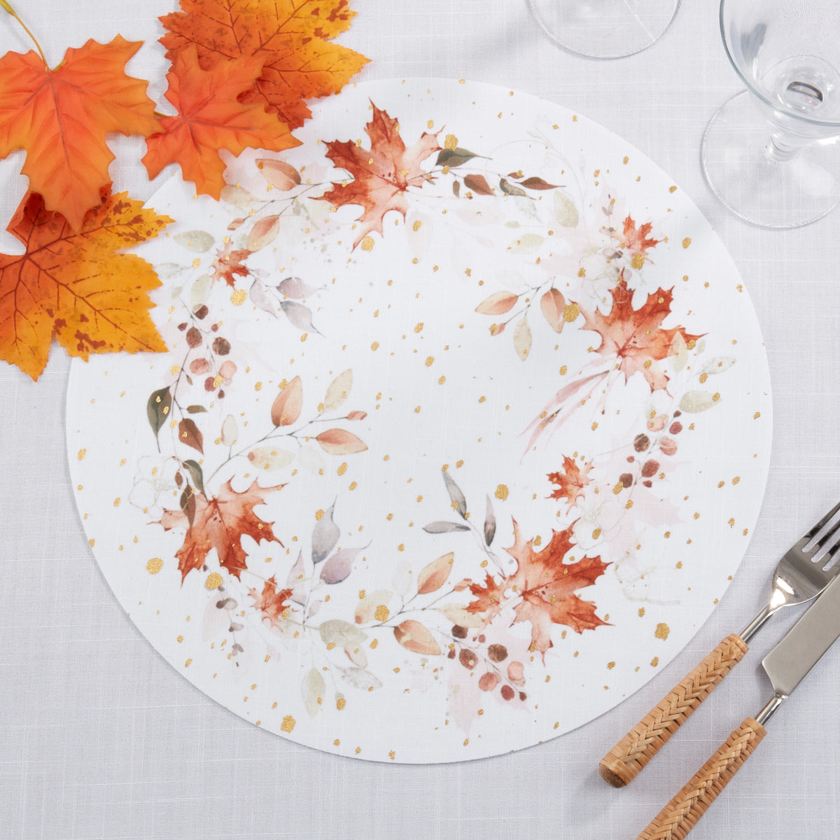 A round white placemat with a watercolor autumn wreath design in orange, red, beige, and muted green, styled on a white tablecloth with fall leaves, glassware, and cutlery.