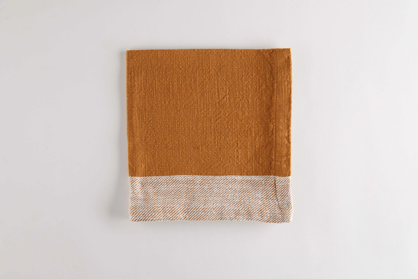 A folded napkin, in warm camel brown with a contrasting woven border in beige and brown, shown on a plain light background.