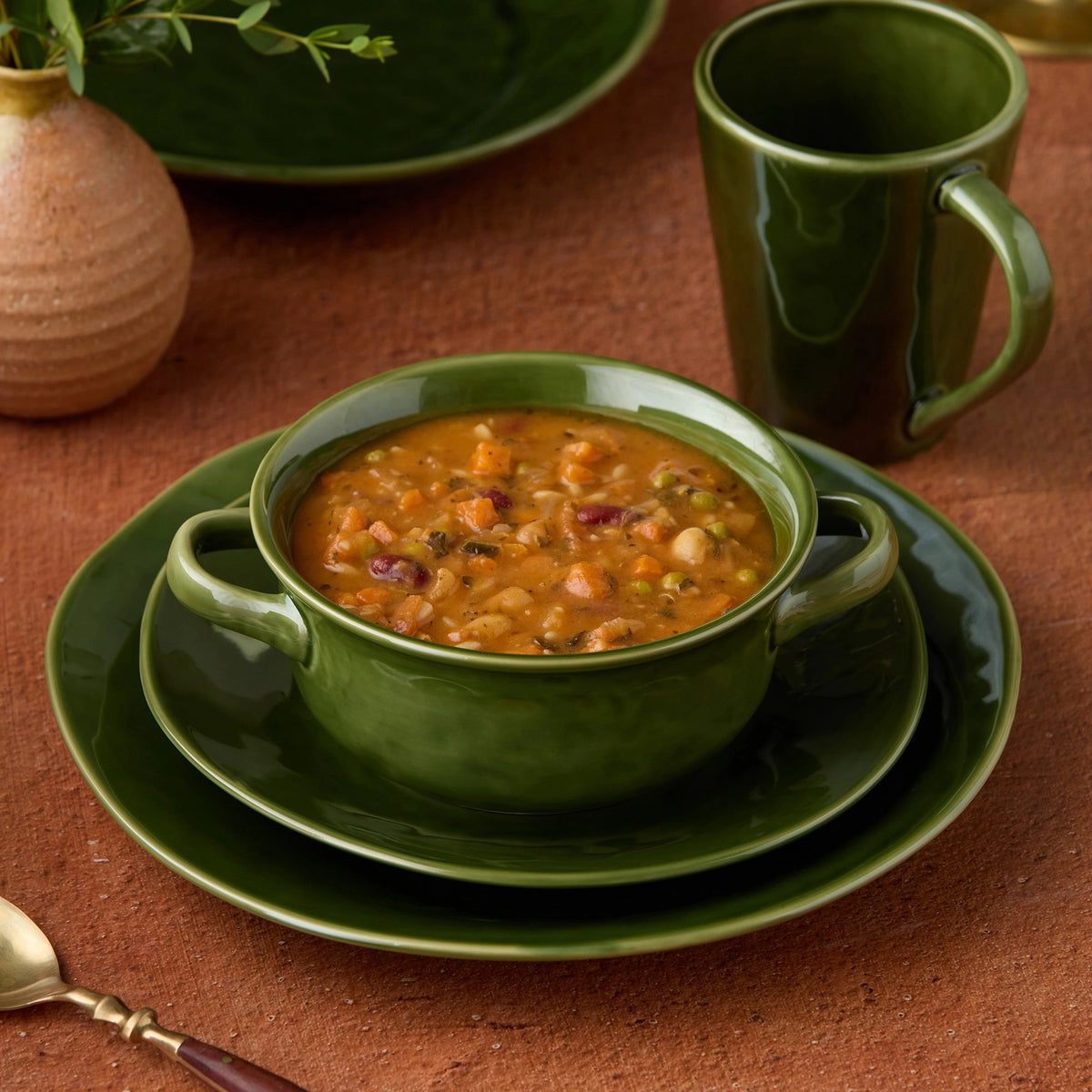 A green ceramic soup bowl filled with chili or bean chili, set on matching green plates, with a green mug and a small vase in a warm brown tabletop setting.