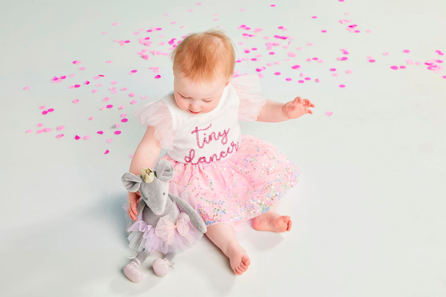 Baby in a 'Tiny Dancer' outfit with a pink sequin tutu, a suffed animal and pink confetti on a light background
