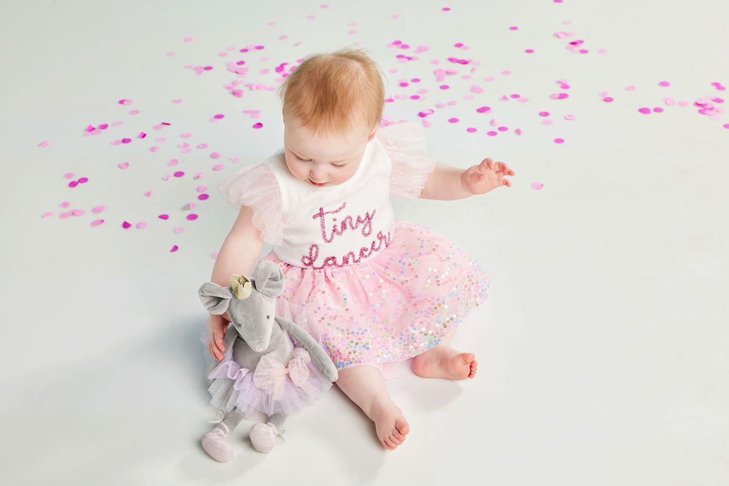 Baby in a 'Tiny Dancer' outfit with a pink sequin tutu, a suffed animal and pink confetti on a light background