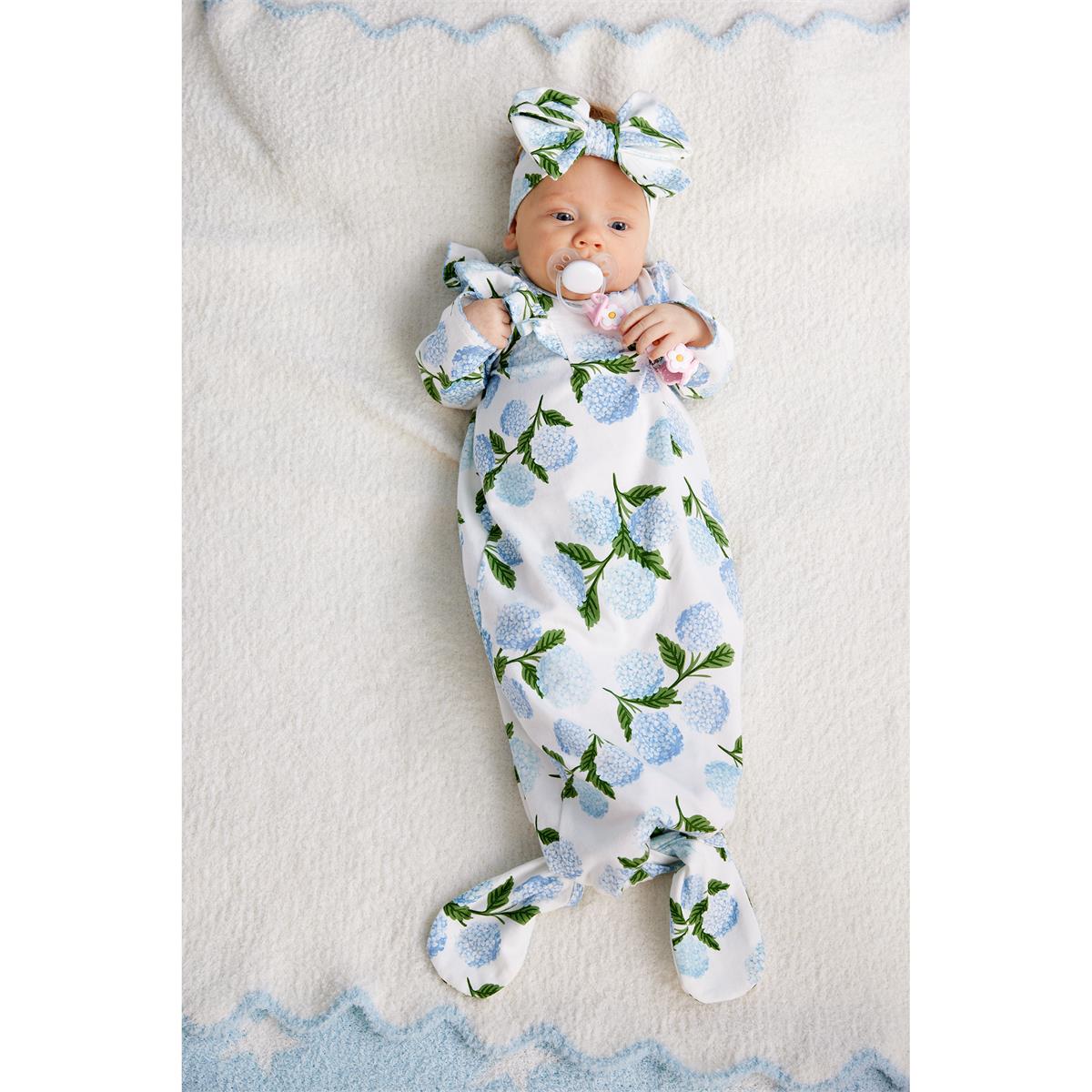 A baby lying on a soft white blanket, wearing a white footed swaddle with a blue and green floral print and a matching bow headband.
