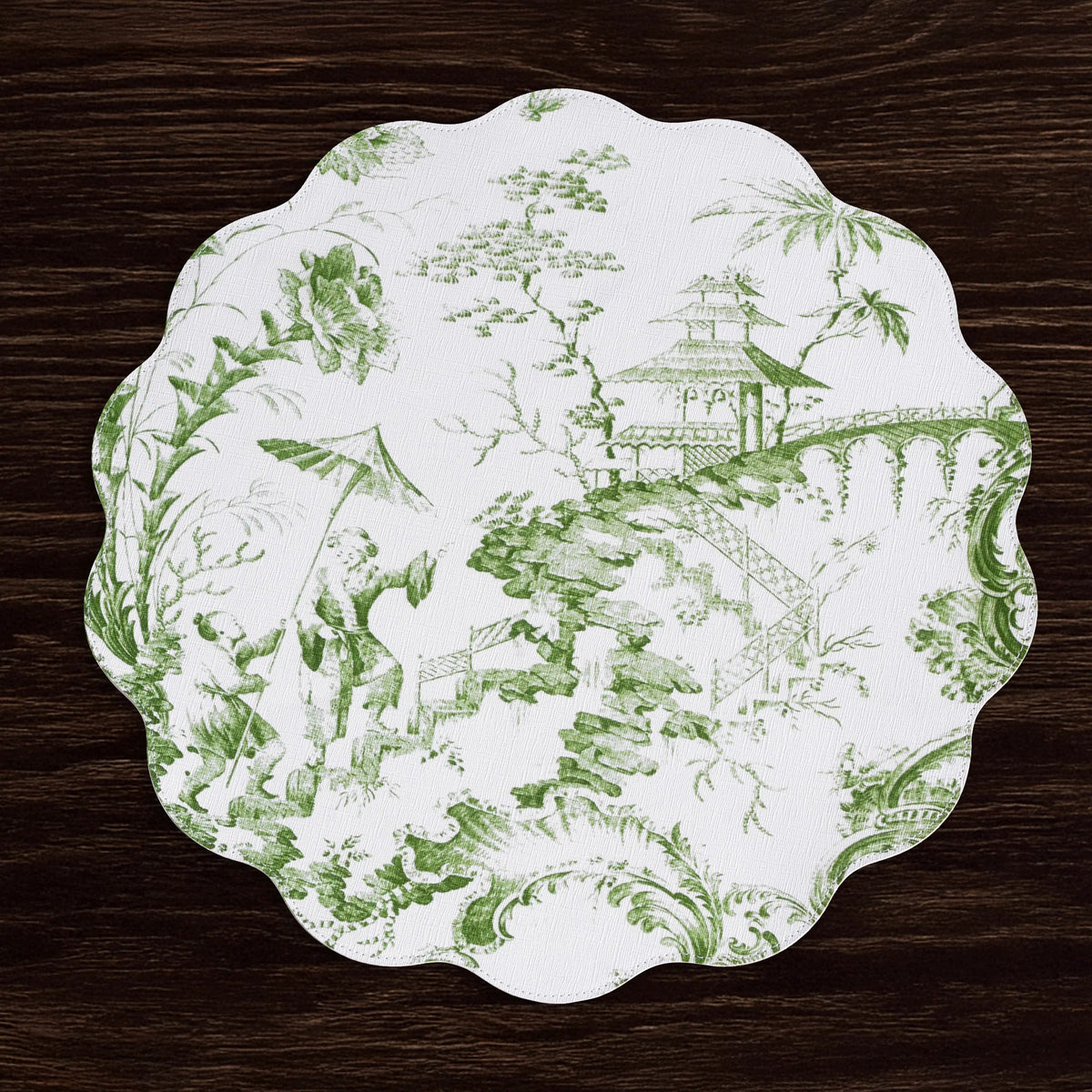A scalloped placemat with a green toile de Jouy landscape pattern on a white background, displayed on a dark wood surface.