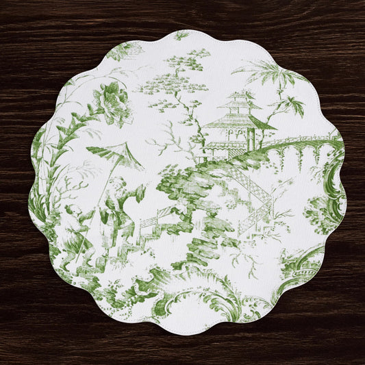 A scalloped placemat with a green toile de Jouy landscape pattern on a white background, displayed on a dark wood surface.