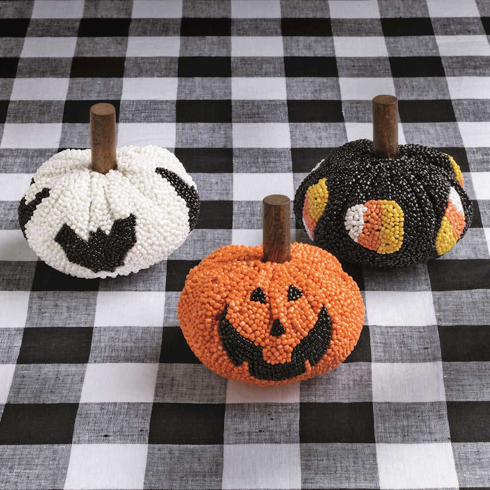 Three handcrafted beaded Halloween pumpkins, displayed on a black and white checkered tablecloth. One pumpkin is white with black bats, one is orange with a black jack-o'-lantern face, and one is black with orange, yellow, and white candy corn details.