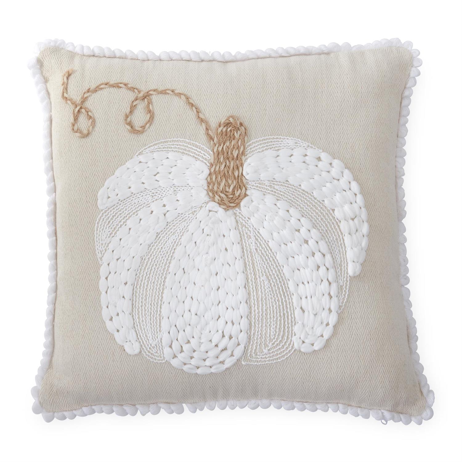 A square beige throw pillow with a white knitted pumpkin design and a tan stem, finished with a white pom-pom border.
