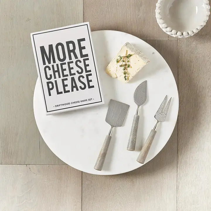 Set of cheese knives with a 'More Cheese Please' sign on a marble surface.