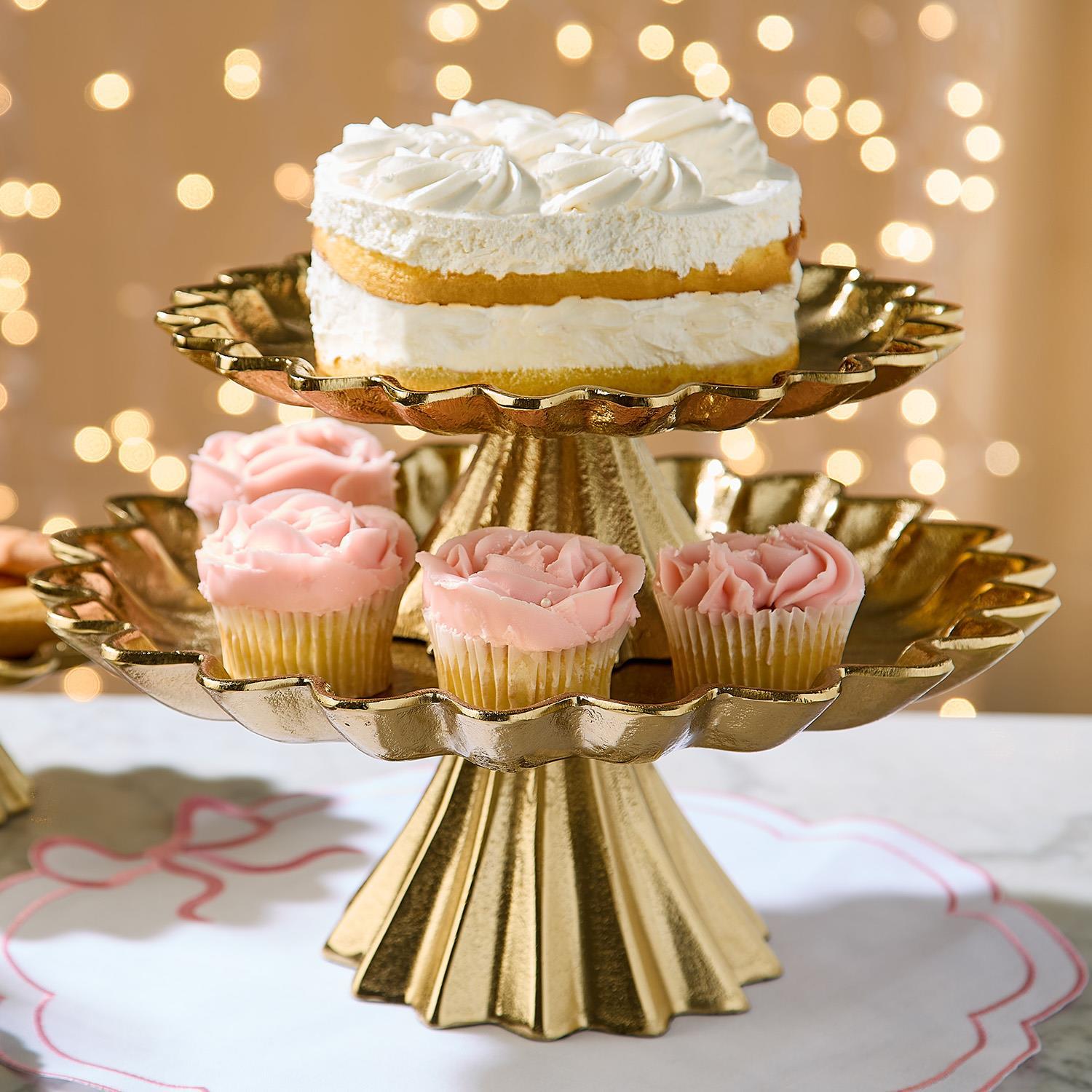Gold cake stand with a layered cake and pink cupcakes against a bokeh light background