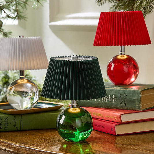 Three pleated table lamps with spherical glass bases in green, red, and clear/white, styled on stacked books on a wooden table.