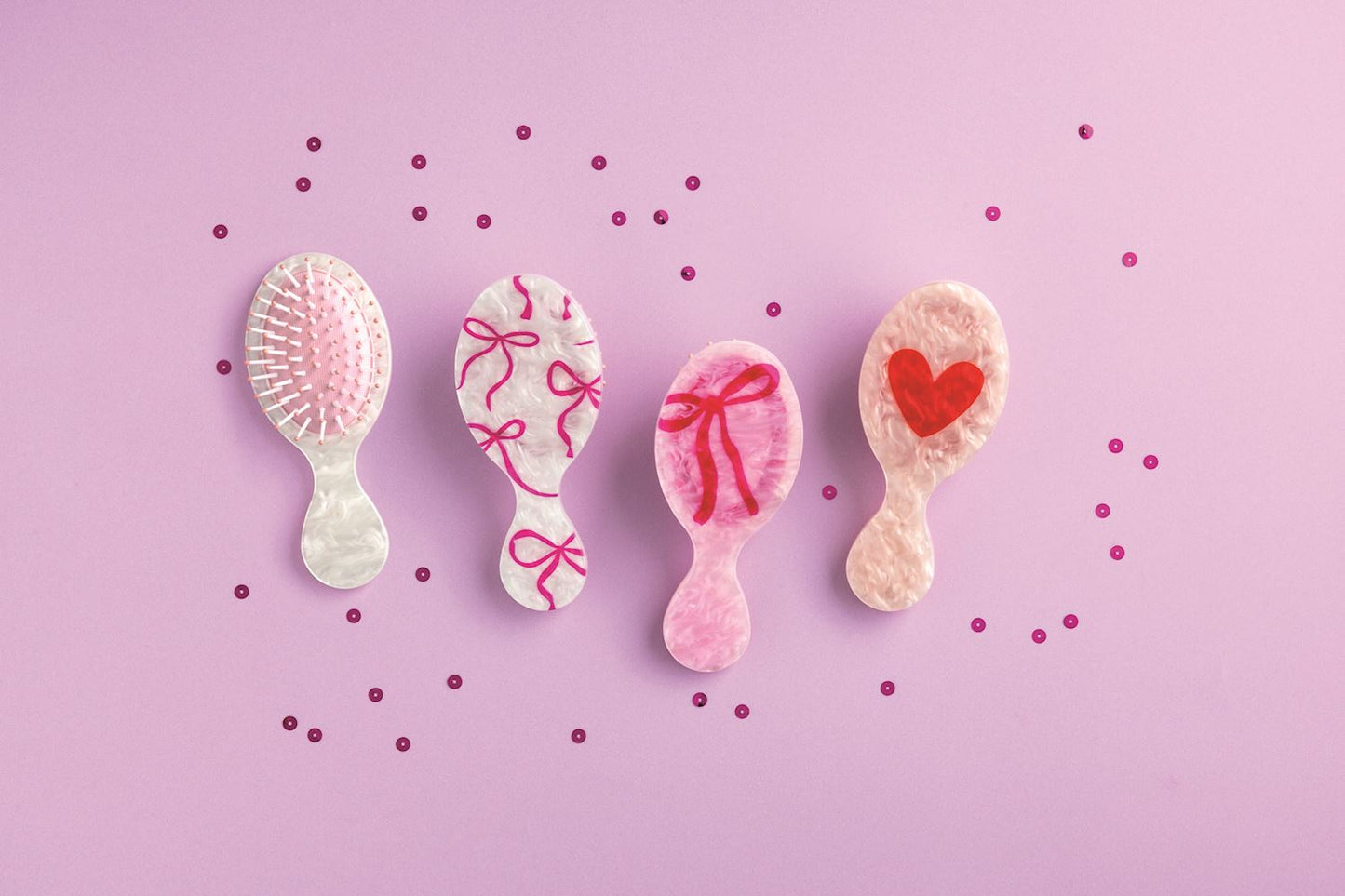 Four Mudpie small hairbrushes with decorative elements on a pink background