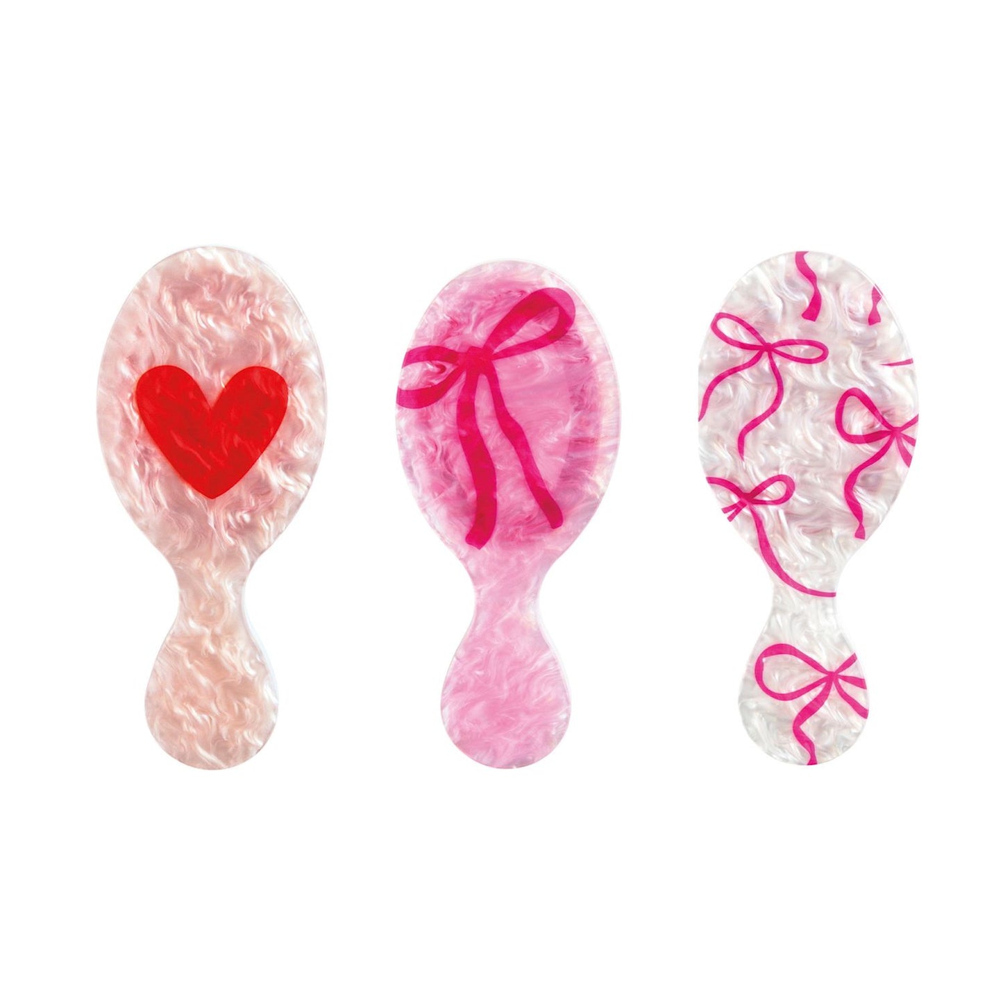 Three decorative Mudpie mini hair brushes in a pearlizedr style, featuring a light pink brush with a red heart, a pink brush with a red bow, and a white brush with pink bows