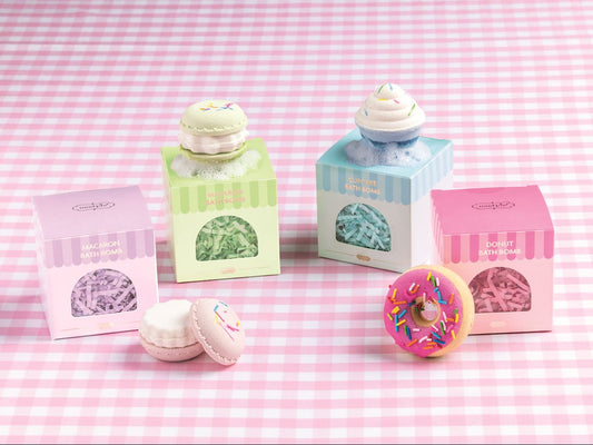 Four small pastel bakery-themed boxes with macarons and a donut toy arranged on a pink-and-white gingham surface. The boxes have striped tops and window fronts showing the contents.