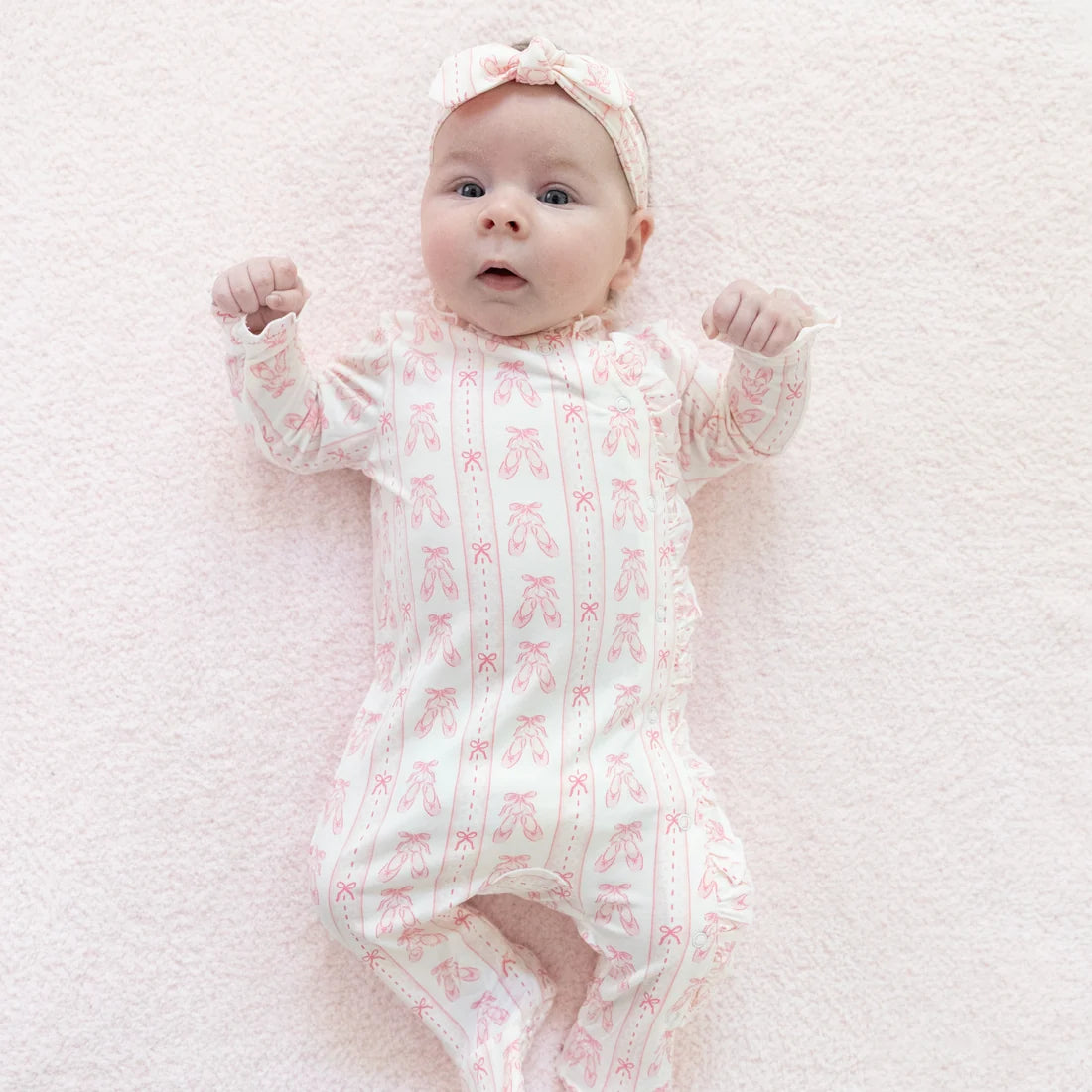 A baby lying on a soft pink blanket, wearing an Angel Dear Ballet Shoes Ribbon Stripe Kimono Ruffle Footie long-sleeve footless onesie with a pink ballet shoe pattern and a matching pink bow headband.