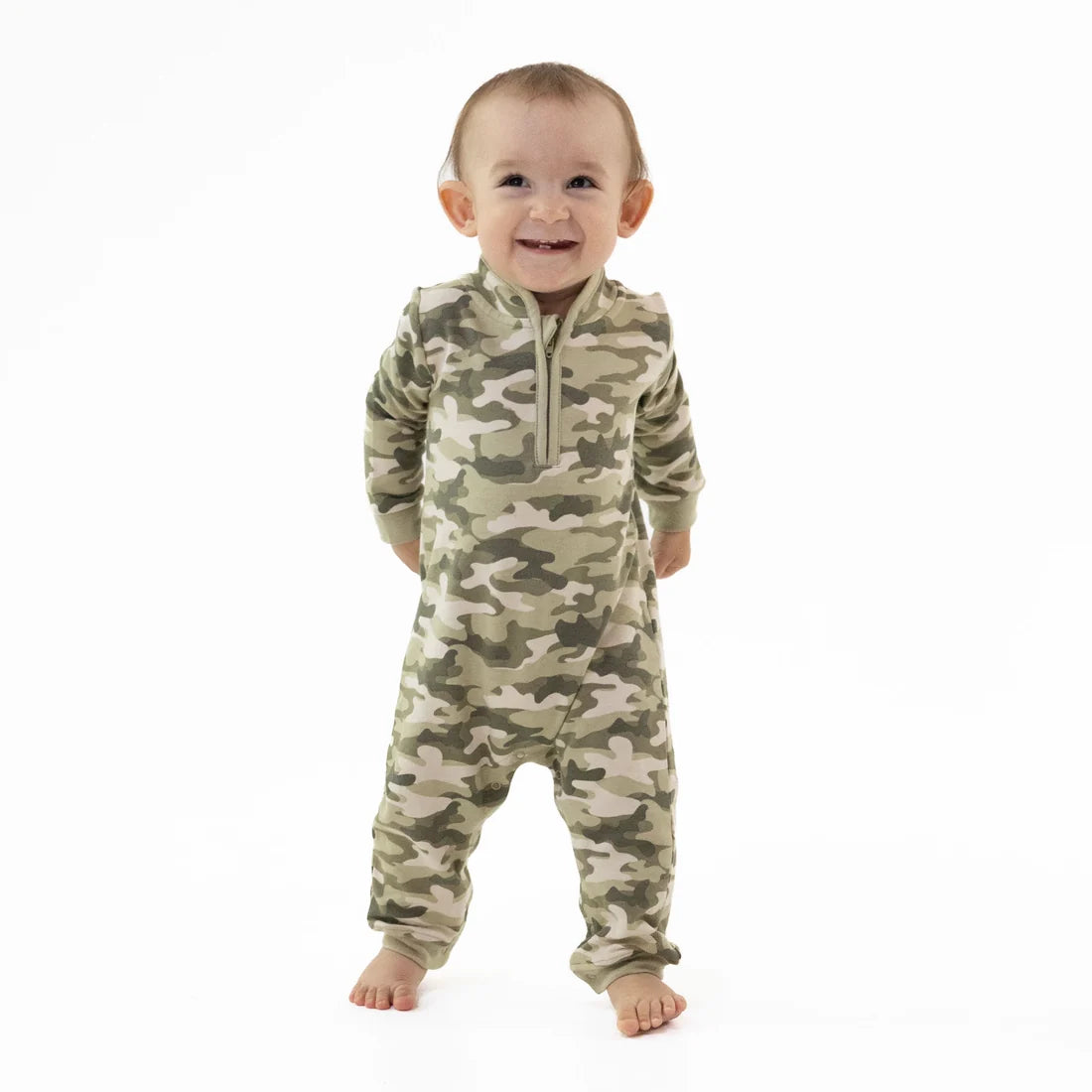 A smiling baby standing barefoot on a white background, wearing a long-sleeve Angel Dear camouflage one-piece french terry romper with a short front zipper.