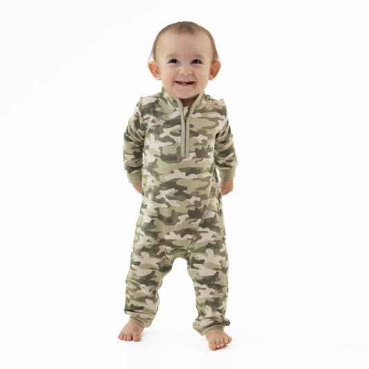 A smiling baby standing barefoot on a white background, wearing a long-sleeve Angel Dear camouflage one-piece french terry romper with a short front zipper.