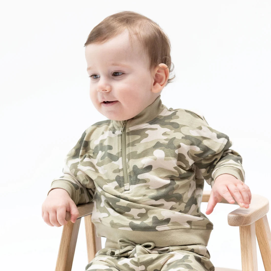 A baby wearing an angel baby long-sleeve camouflage half-zip romper sits on a small wooden chair against a plain white background.