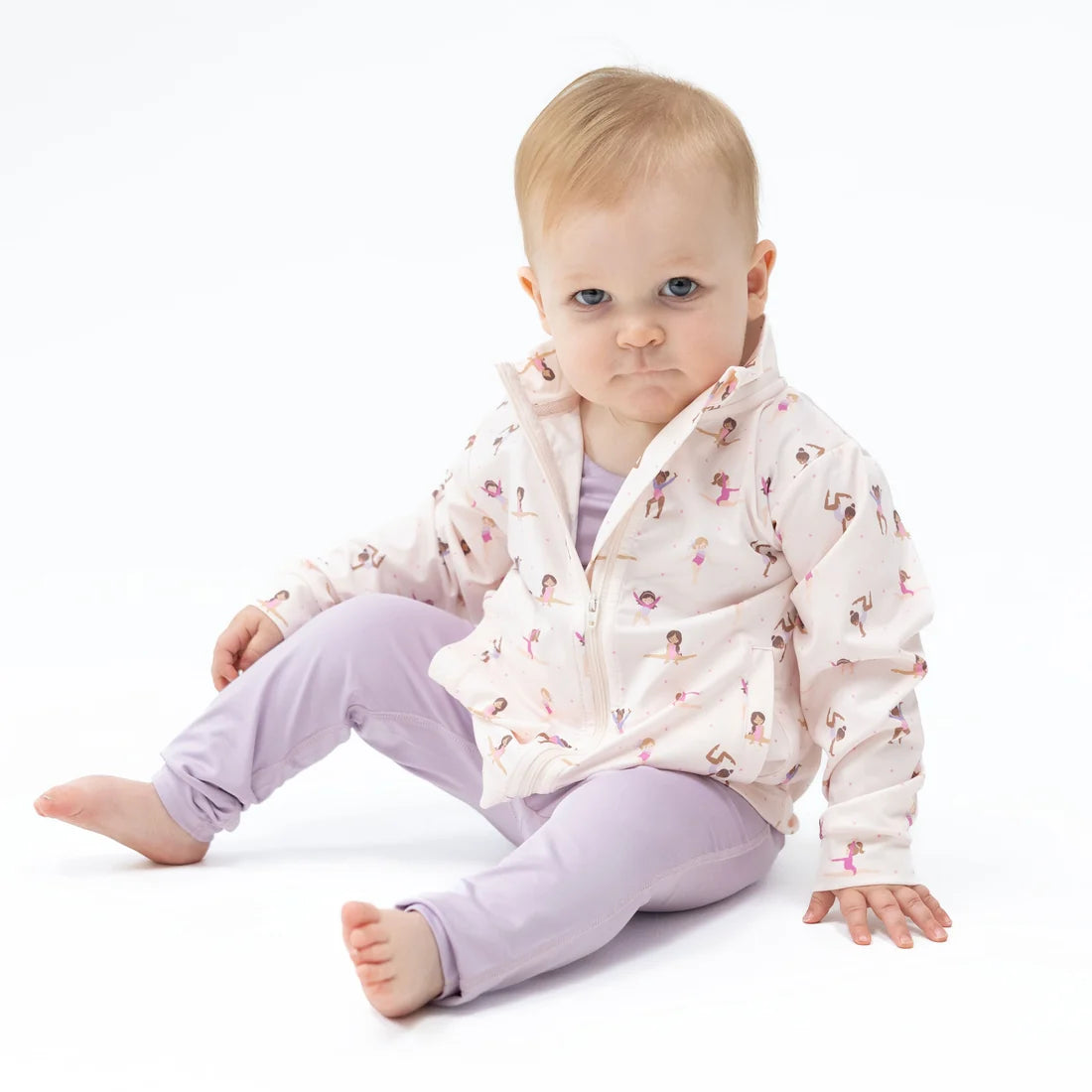 Baby wearing a lavender romper with an Angel dear tiny tumblers white jacket with images of girls doing tumbliing activities sitting on a studio floor with a white background
