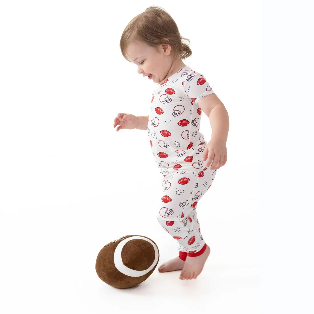 A toddler standing barefoot on a white studio background, wearing a white  Angel dear touchdown time short-sleeve one-piece pajama with a red and black football pattern, next to a plush football toy.