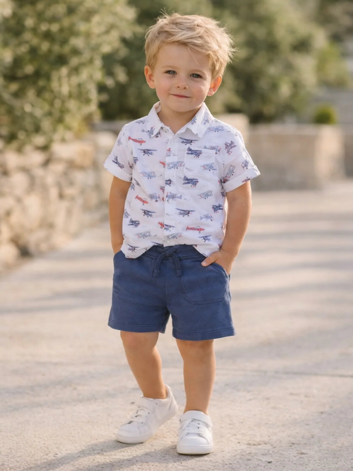 Young boy wearing an angel dear vintage airplane patterned shirt and blue shorts standing outdoors.