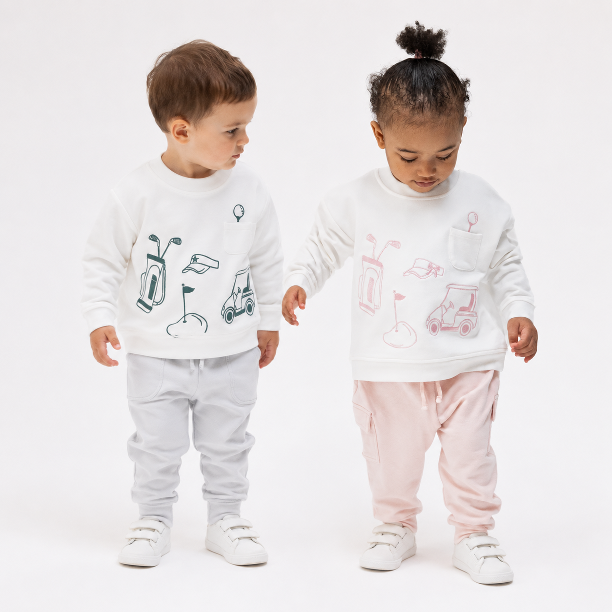 Two toddlers standing side by side in a studio setting, wearingAngel dear matching white long-sleeve sweatshirts with golf-themed graphics. The boy wears light gray joggers and white sneakers, while the girl wears light pink joggers and white sneakers.