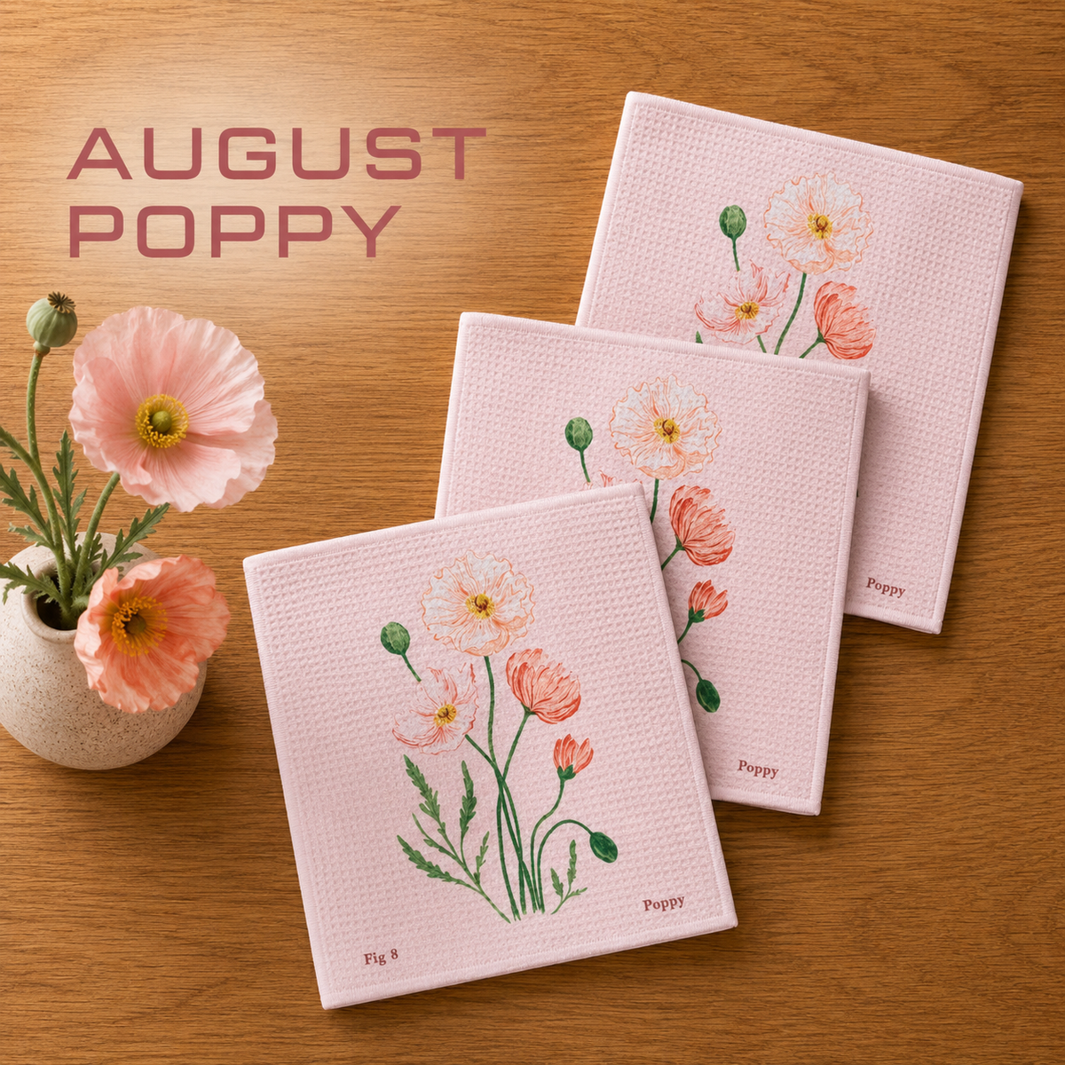 Geometry August Poppy Dishcloth set with Three textured pink square dishcloths featuring a watercolor-style poppy illustration, arranged on a wooden surface next to a small vase of pink flowers. The text 'AUGUST POPPY' appears in the upper left.  The Little Exchange SKU 141323