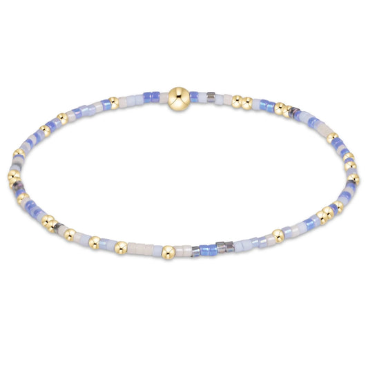A delicate beaded enewton hope unwritten bloopers bracelet with white and  light blue and white translucent beads, accented by shiny gold metallic beads and a central gold clasp, shown on a plain white background.