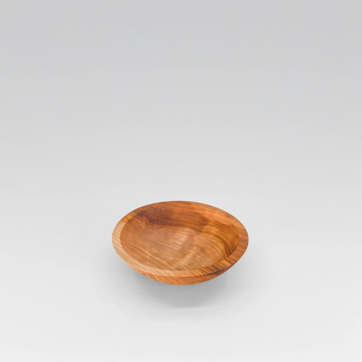 A small, round cherry wooden bowl by holland bowl mill with a warm medium-brown finish and visible wood grain, centered on a light gray gradient background.