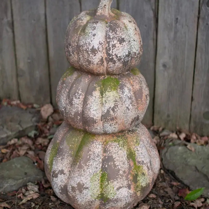 A three-tiered decorative pumpkin sculpture with a weathered, mossy finish, standing outdoors against a wooden fence.