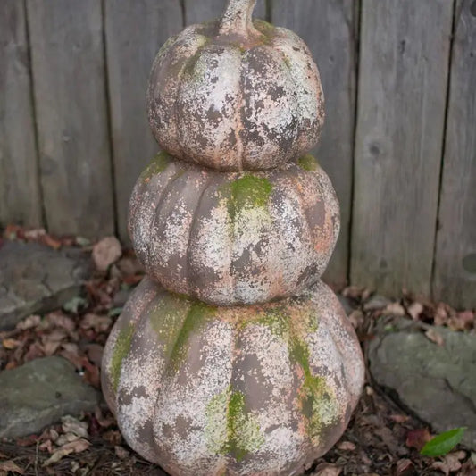 A three-tiered decorative pumpkin sculpture with a weathered, mossy finish, standing outdoors against a wooden fence.