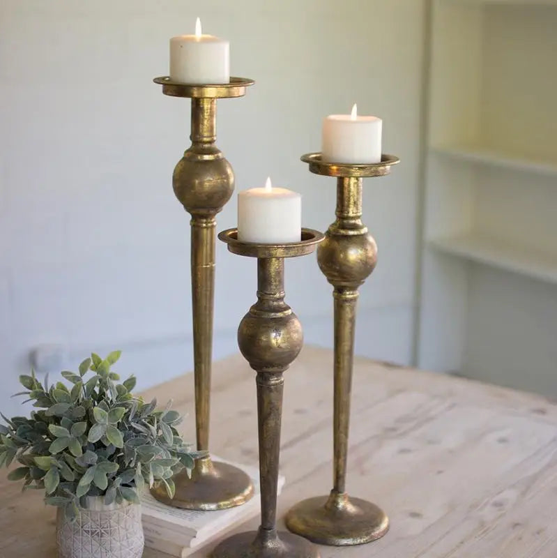 Three vintage-style antique brass candlestick holders with white pillar candles, styled on a light wooden table next to a small green plant.