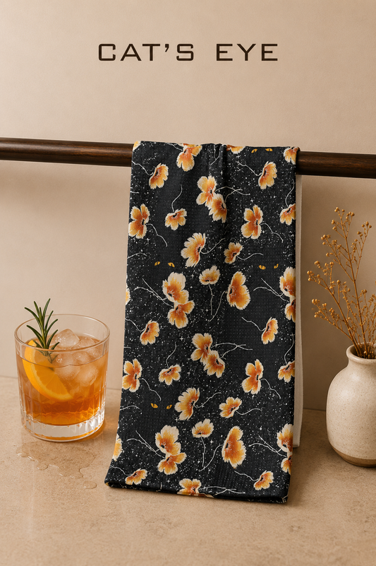 A black Geometry Cat's Eye bar towel with a yellow and orange floral pattern hangs from a dark wooden rod. The scene is styled with a cocktail in a glass on the left and a small vase of dried stems on the right, against a beige wall with the text 'CAT'S EYE' at the top.  The Little exchange SKU 150067