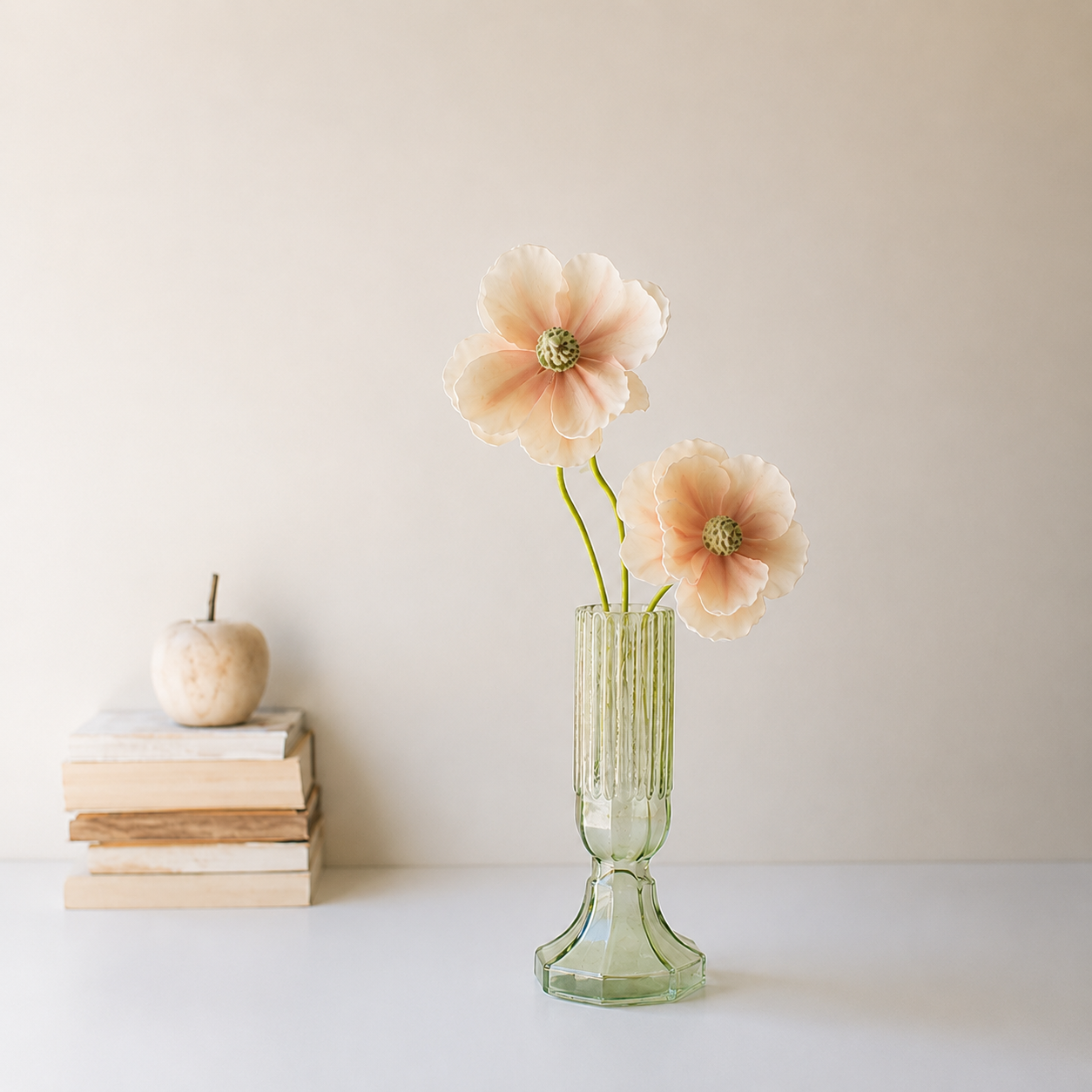 A light green ribbed Kalalou glass vase with a flared base holding two pale peach artificial flowers, styled on a white surface against a warm beige wall with stacked books and a small decorative object in the background.
