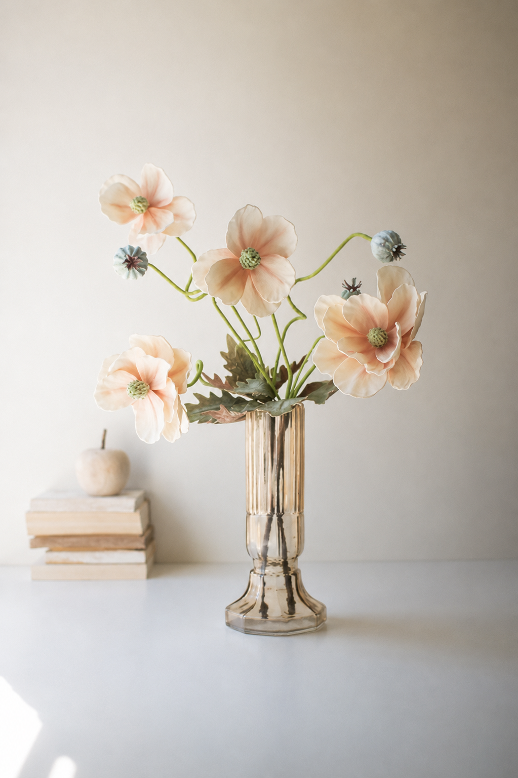 A vase of artificial peach anemone flowers with green stems and blue-gray buds, displayed in a ribbed clear Kalalou yellow glass vase on a white surface next to a stack of books and a small decorative pumpkin against a neutral beige background.
