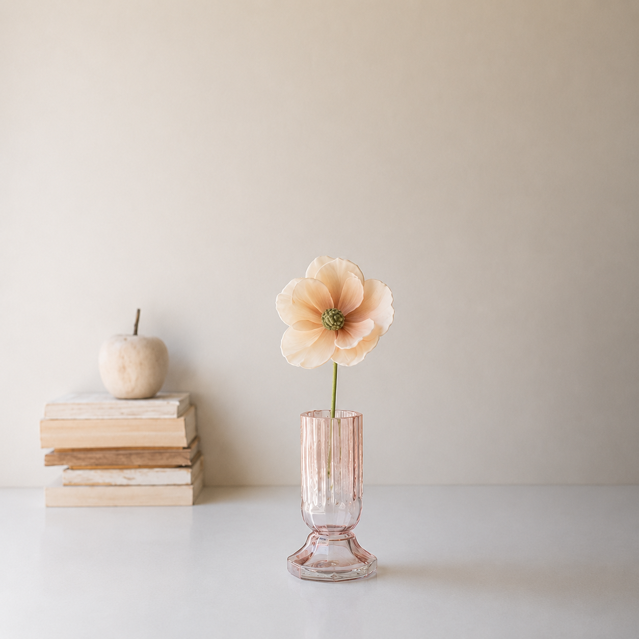 A pale peach artificial flower in a ribbed, translucent Kalalou glass vase, styled on a white surface next to a stack of books and a small wooden apple against a warm beige wall.