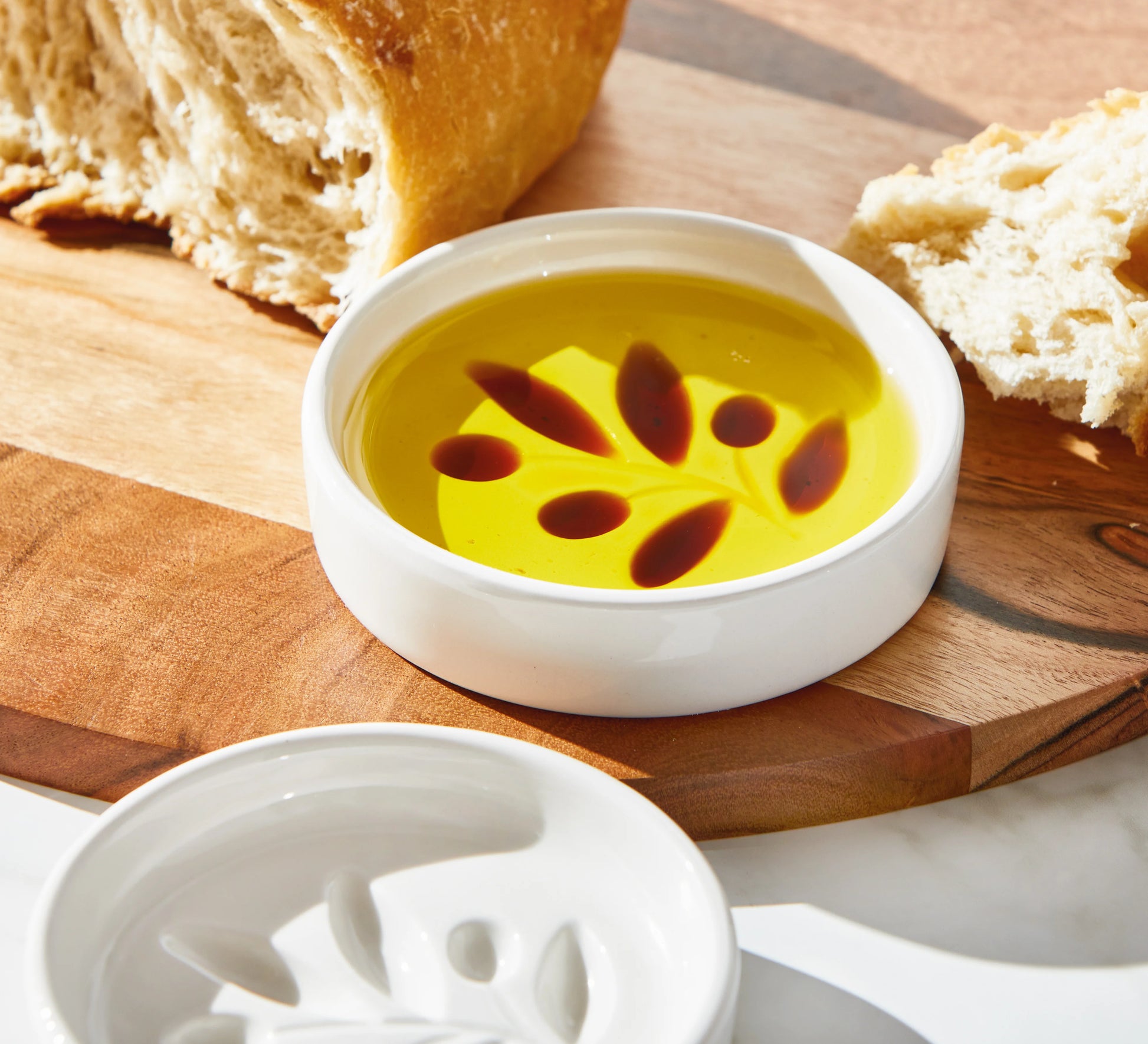 Small bowl of oil and vinegar in a tag olive branch dipping dish on a wooden board with bread slices.
