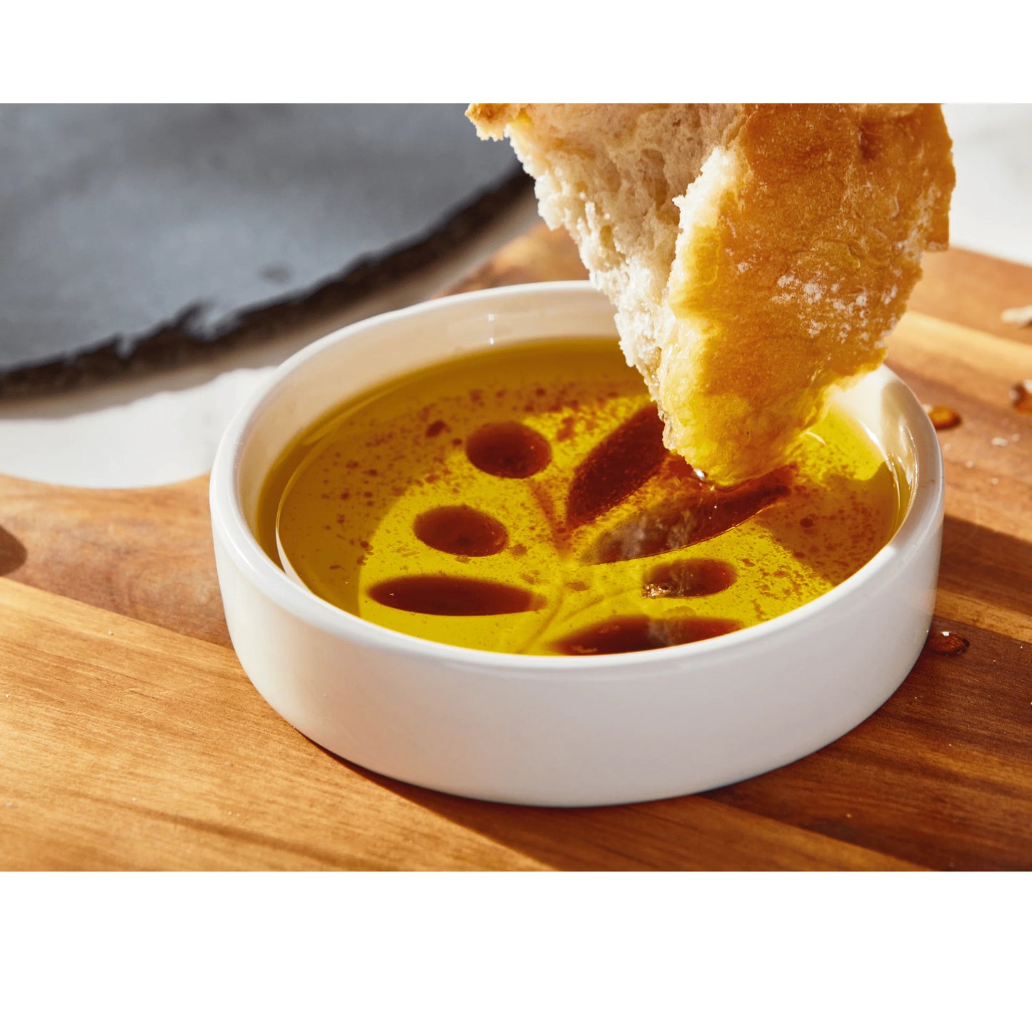 Dipping bread into a bowl of olive oil in a white oil and vinegar tag dipping dish on a wooden surface