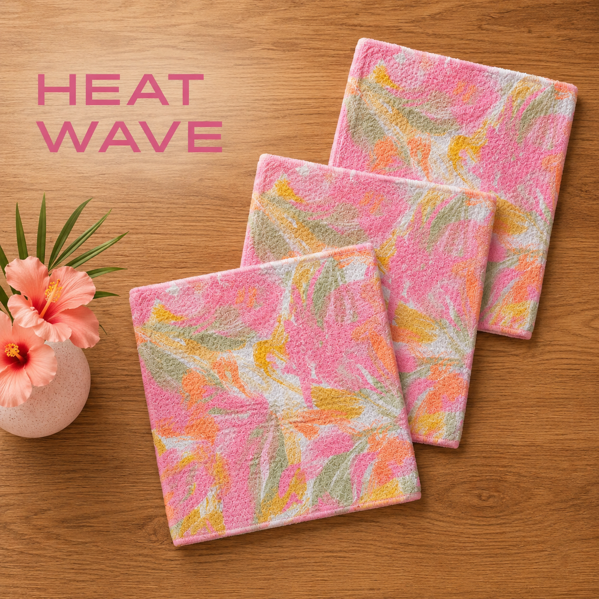 Geometry Heat Wave dischcloth set with Three square pink Heat Wave plush bath towels with a tropical leaf pattern in pink, yellow, and green, arranged on a wooden surface next to a small vase of hibiscus flowers. The text 'HEAT WAVE' is displayed in the upper left corner.  The Little Exchange SKU 142491