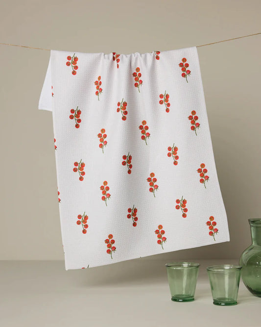 White Geometry tea towel with red tomato pattern hanging on a line, with green glasses and a vase below.