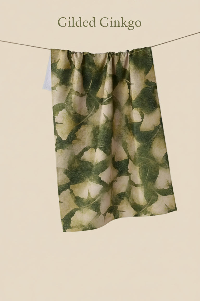 A Geometry GIlded Ginkgo tea towel l hanging from a thin wire with  an allover botanical leaf pattern in dark green and warm beige/cream tones.