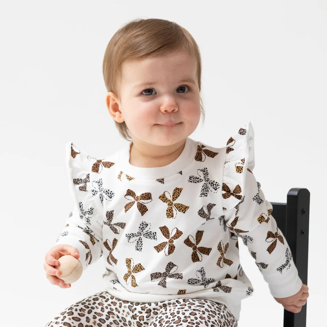 A baby wearing  Angel dear leopard print sweatshirt and leggings 
a white long-sleeve top with brown bow and butterfly patterns, paired with leopard-print pants, sitting on a black chair against a white background.